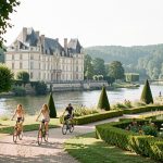 Visiter la vallée de la Loire : 8 expériences mémorables Vue lumineuse d’un château de la Loire au bord d’une rivière, entouré de jardins français, avec quatre cyclistes sur un chemin arboré.